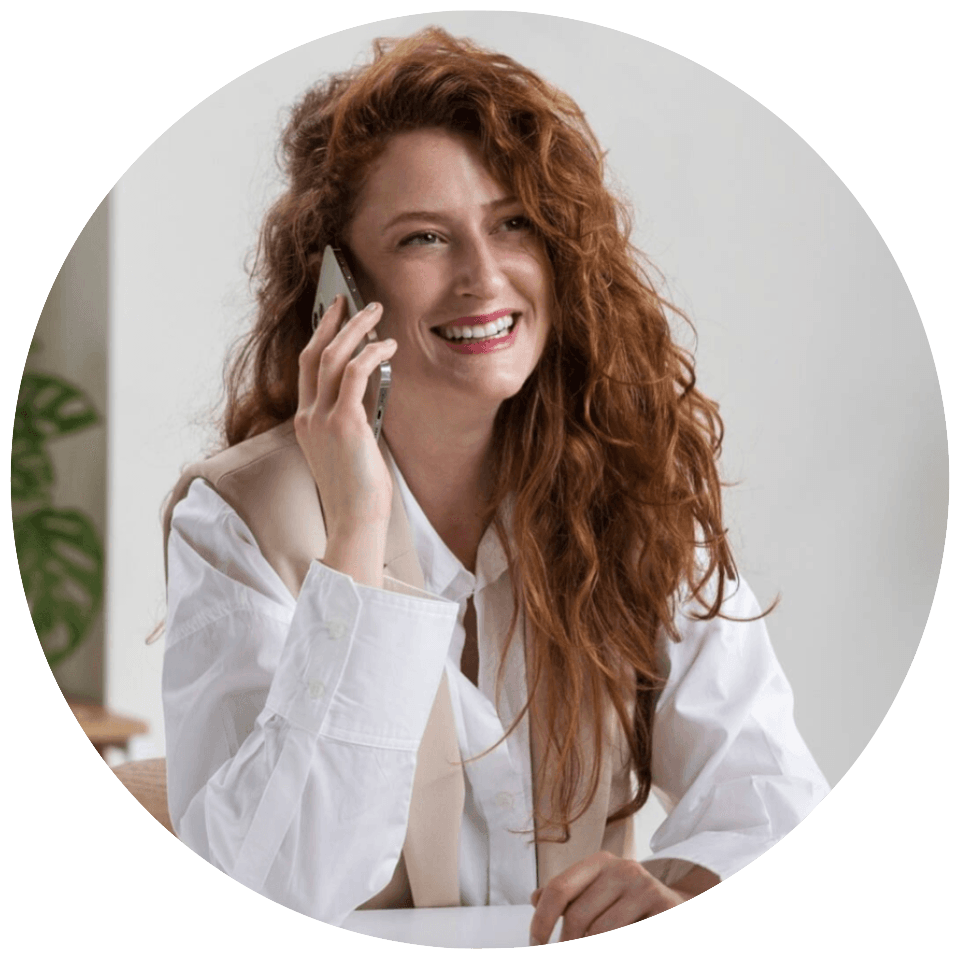 "Femme rousse souriante en pleine conversation téléphonique assise à son bureau"