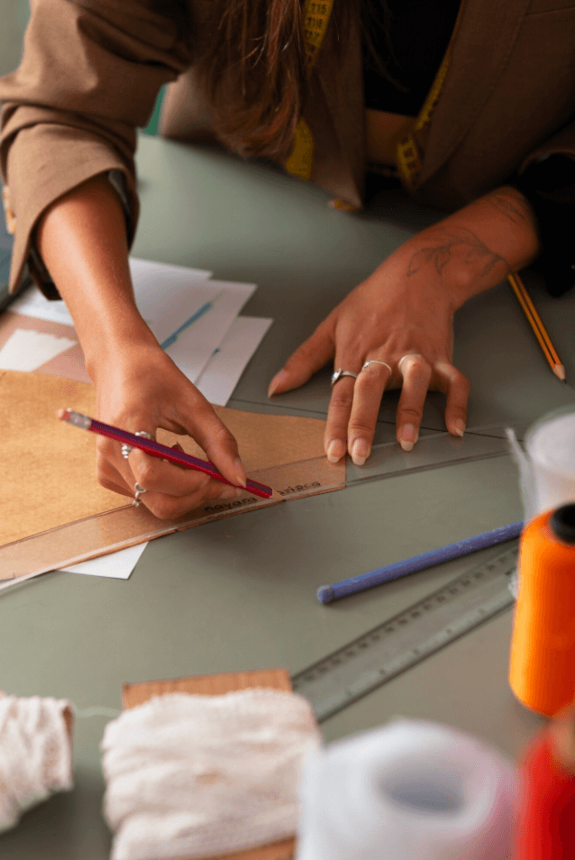 alt="Gros plan sur les mains d'une créatrice traçant une ligne sur un patron de couture avec une règle et un crayon, entourée de bobines de fil colorées"