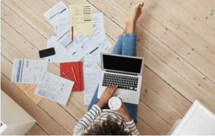 alt="Vue de dessus d'une personne assise à même le sol travaillant sur un ordinateur portable, entourée de nombreux documents administratifs éparpillés, d'un carnet rouge et tenant un café"