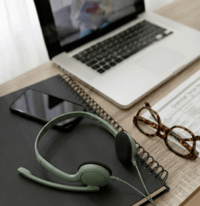 alt="Ordinateur portable ouvert sur un bureau affichant une visioconférence, avec un casque audio vert, des lunettes et un carnet au premier plan"