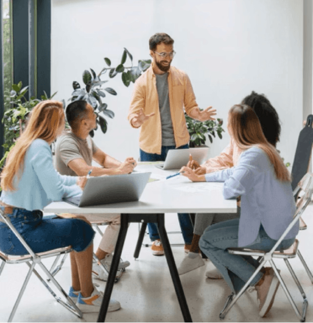 alt="Un homme debout prend la parole devant quatre participants à atelier assis autour d'une table blanche dans un bureau moderne et lumineux décoré de plantes"