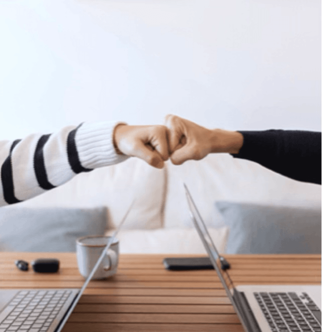 alt="Deux personnes se font un check du poing au-dessus d'une table en bois où sont posés deux ordinateurs portables ouverts face à face"