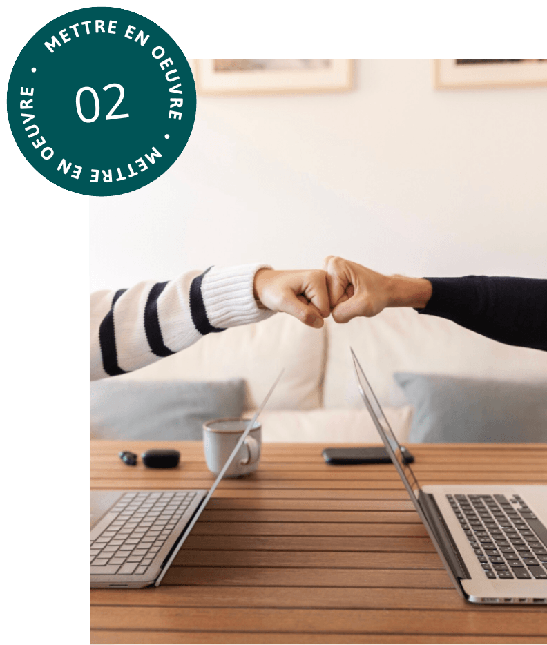 alt="Deux personnes se font un check du poing au-dessus d'une table en bois où sont posés deux ordinateurs portables ouverts face à face"