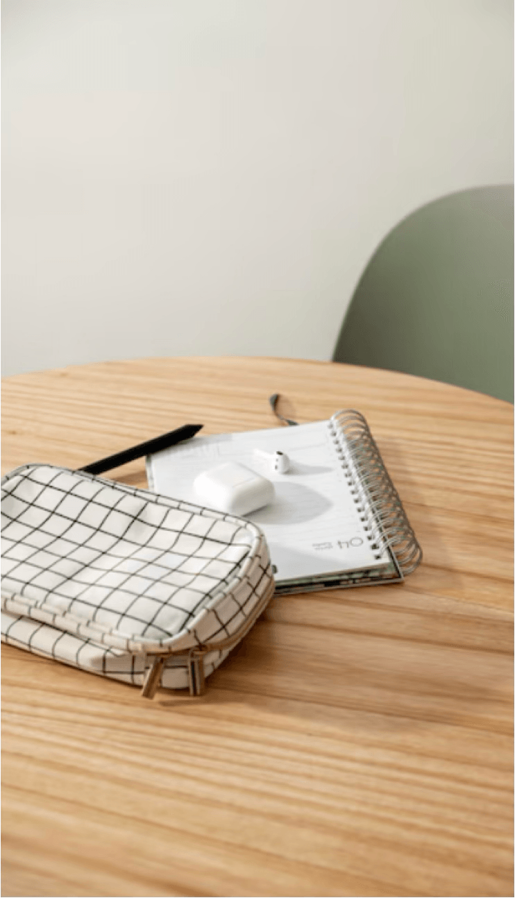 alt="Trousse à motifs quadrillés, carnet à spirales ouvert et écouteurs sans fil posés sur une table ronde en bois clair"