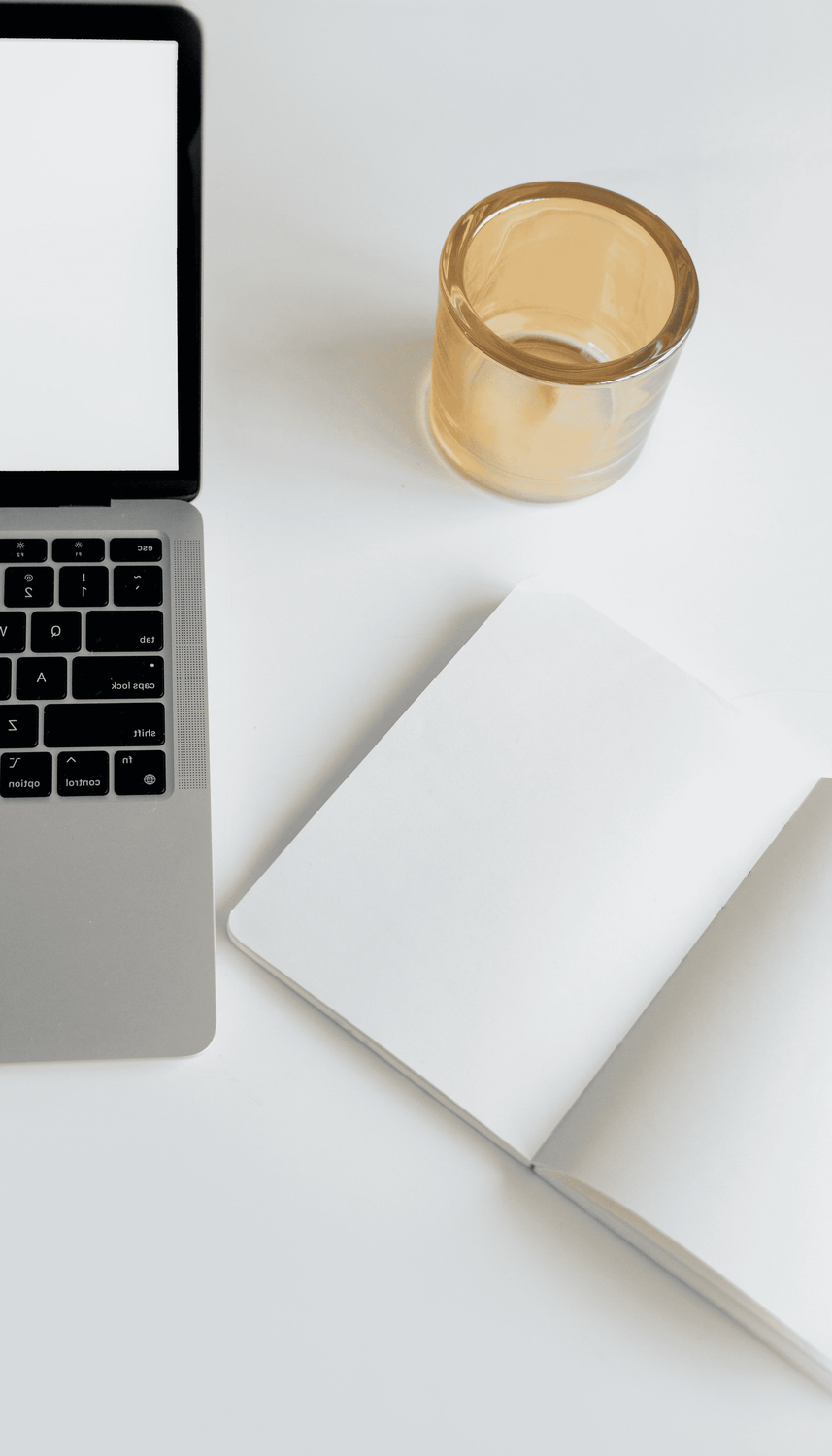 alt="Vue de dessus d'un bureau blanc avec un ordinateur portable, un carnet ouvert aux pages blanches et un verre ambré"