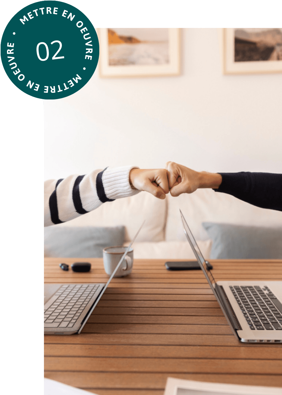 alt="Deux personnes se font un check du poing au-dessus d'une table en bois où sont posés deux ordinateurs portables ouverts face à face"