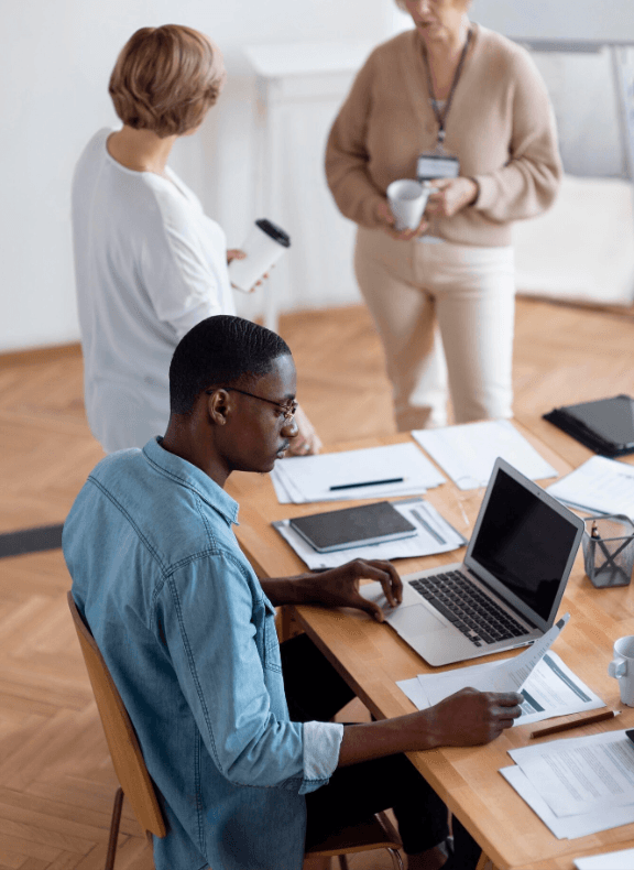 alt="Au sein d'une collectivité, un homme à lunettes travaille sur son ordinateur portable, tandis que deux femmes discutent debout en arrière-plan avec des tasses de café"