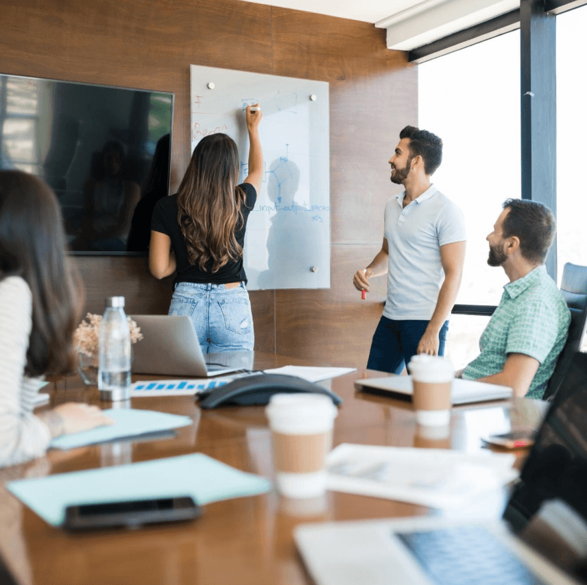 alt="Une femme écrit sur un tableau blanc lors d'une formation, tandis qu'un collègue debout sourit et que d'autres participants sont assis autour d'une table de conférence"