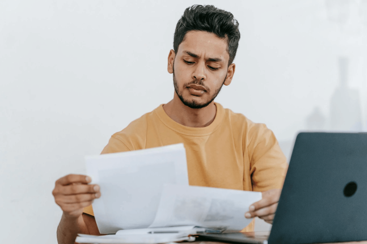 alt="Un homme concentré lit attentivement des documents imprimés qu'il tient à deux mains, assis devant son ordinateur portable ouvert"