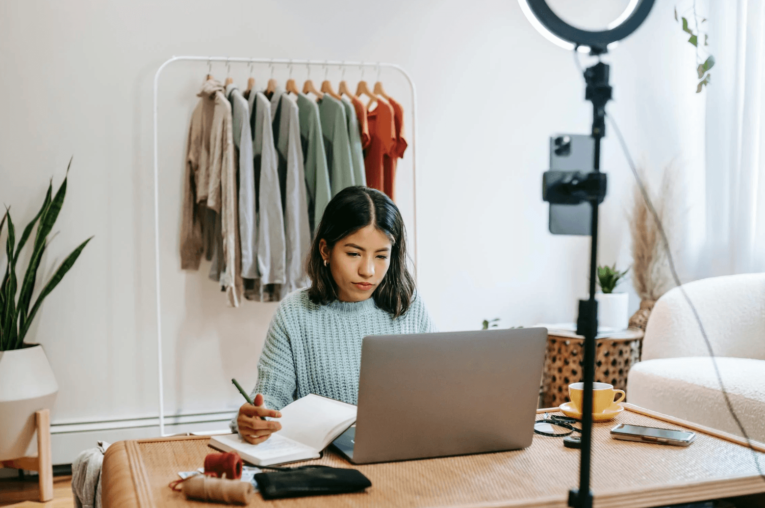 alt="Créatrice solopreneure prend des notes dans un carnet devant son ordinateur portable, pour préparer sa communication vidéo ou la mise en ligne de ses collections depuis son espace de travail à domicile"