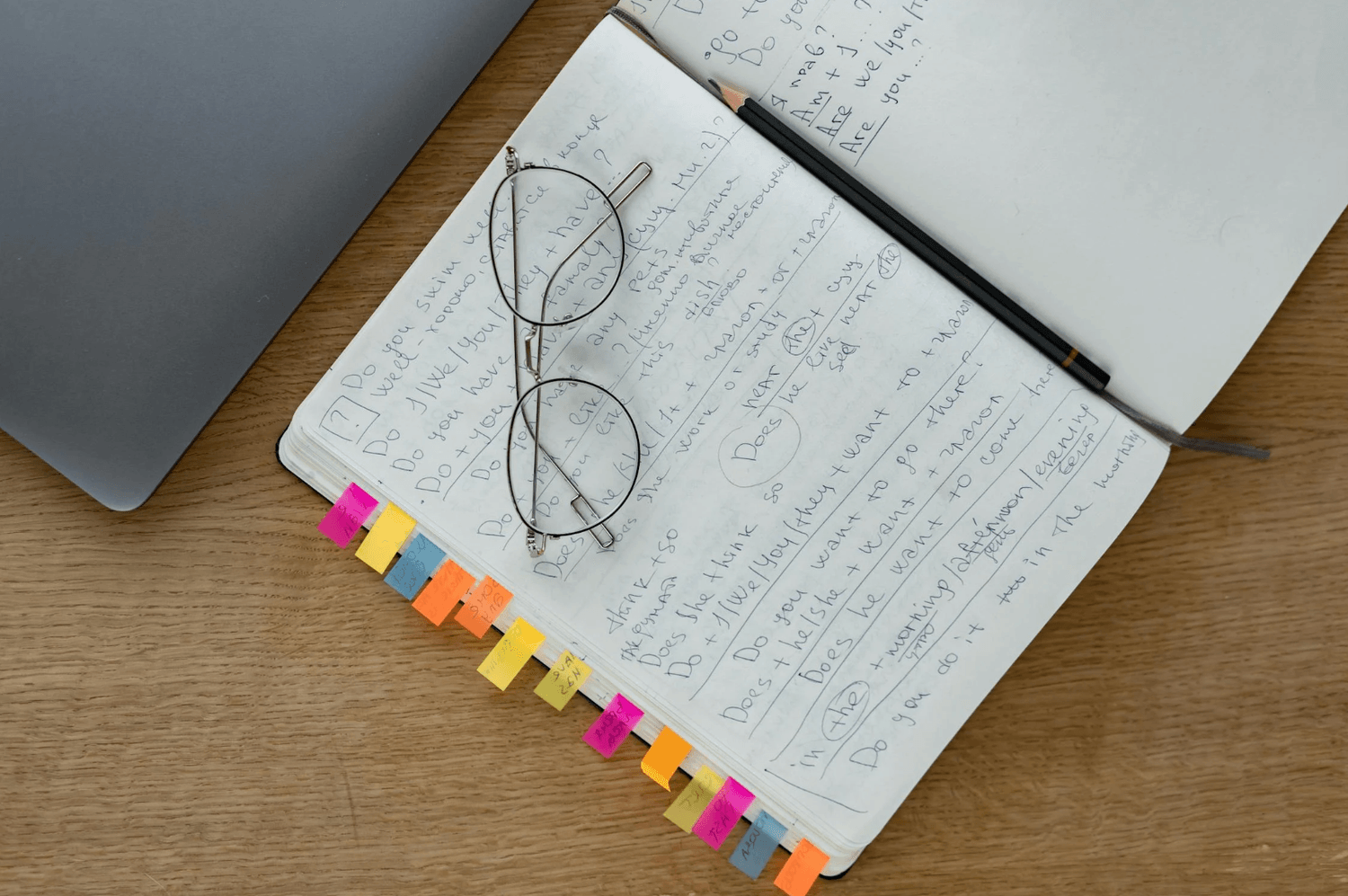 alt="Vue de dessus d'un carnet ouvert rempli de notes manuscrites et de nombreux onglets colorés, surmonté d'une paire de lunettes rondes et d'un crayon, à côté d'un ordinateur portable"