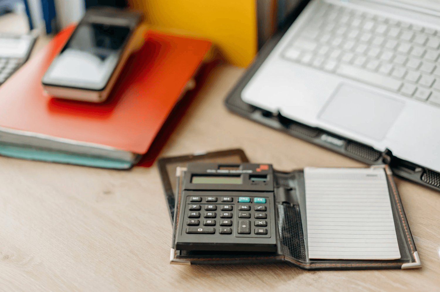 alt="Gros plan sur un bureau en bois avec une calculatrice et un bloc-notes au premier plan, devant un ordinateur portable et des dossiers colorés en arrière-plan"