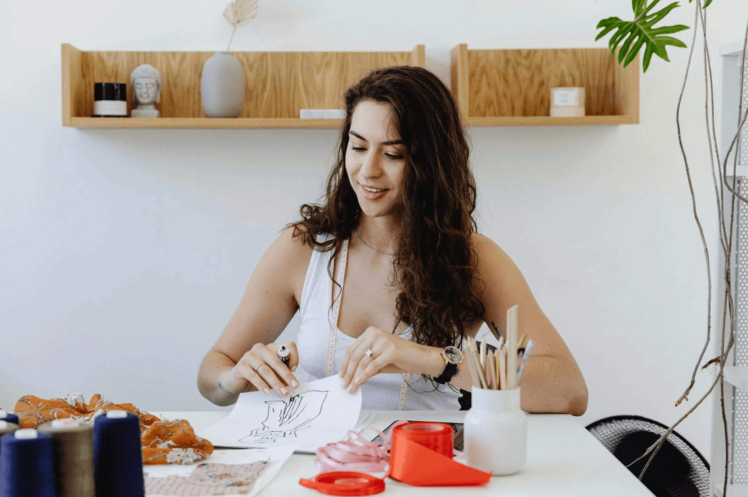 alt="Une créatrice souriante assise à un bureau blanc, travaillant sur un croquis de mode, entourée de bobines de fil, de rubans et de tissus"