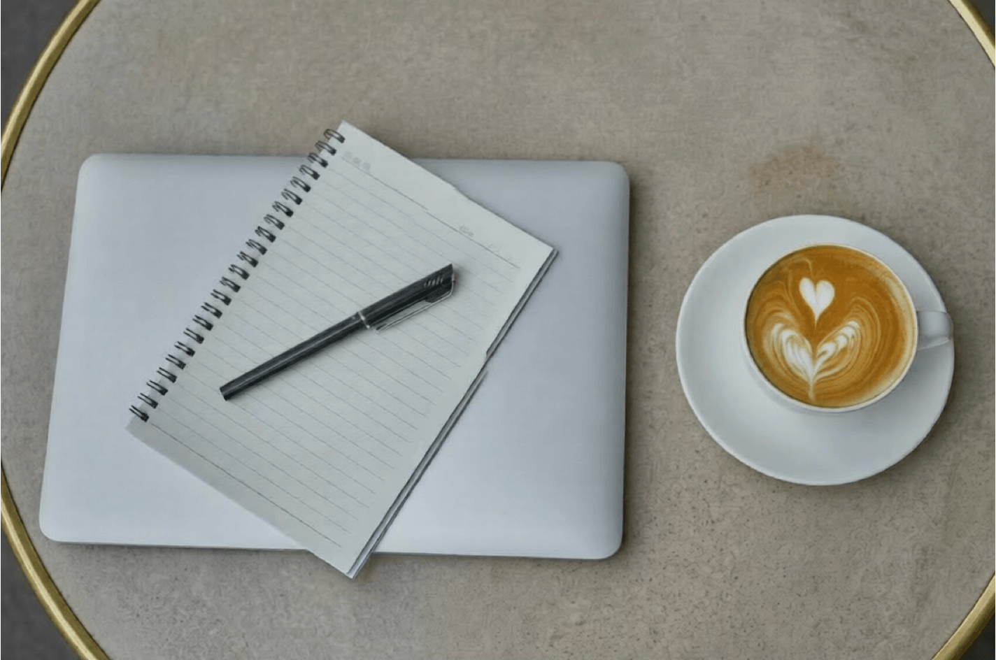 alt="Vue de dessus d'une table ronde avec un ordinateur portable fermé, un carnet à spirales, un stylo noir et une tasse de café avec un motif en forme de cœur"