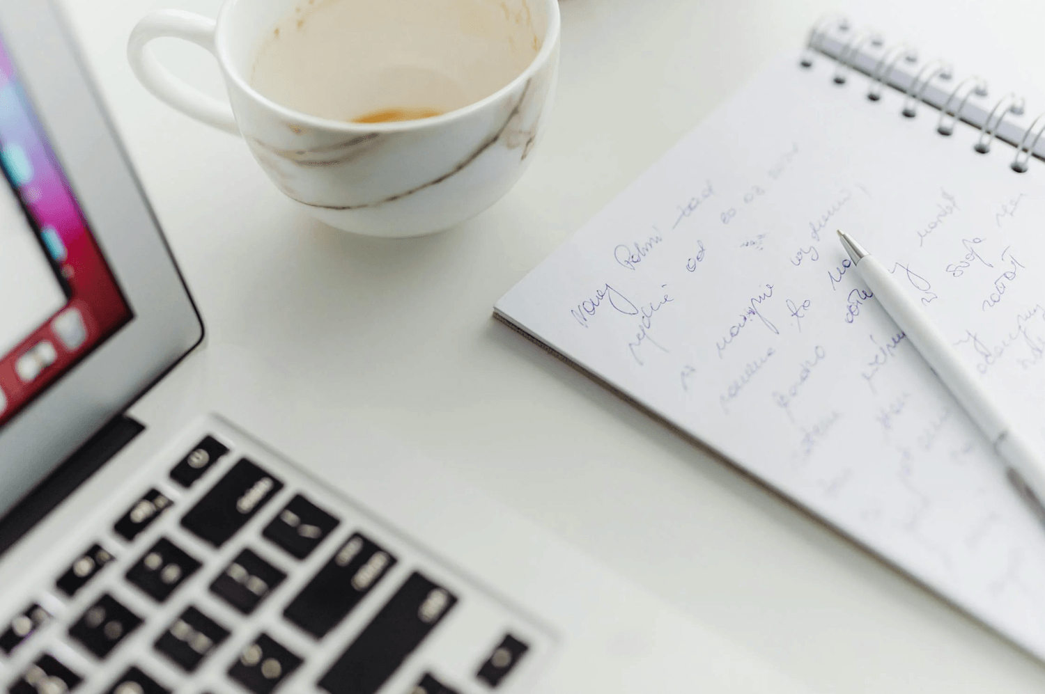 alt="Gros plan sur un bureau blanc avec un coin d'ordinateur portable, une tasse de café presque vide et un carnet à spirales rempli de notes manuscrites avec un stylo blanc"