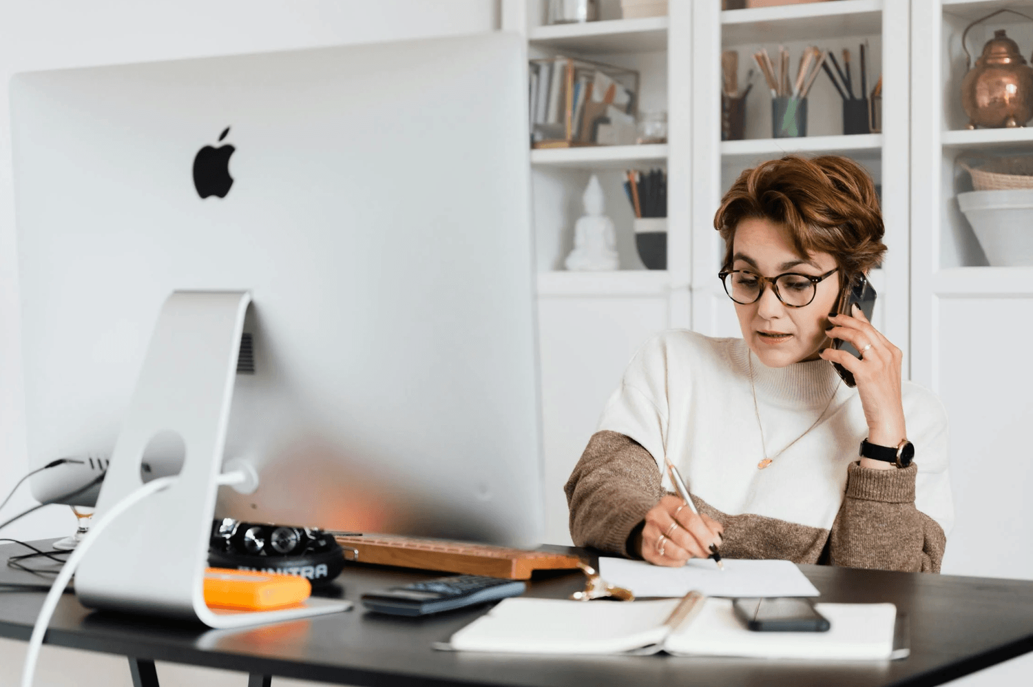 alt="Une femme à lunettes d'une cinquantaine d'années assise à son bureau devant un grand écran d'ordinateur, prenant des notes manuscrites tout en discutant au téléphone"
