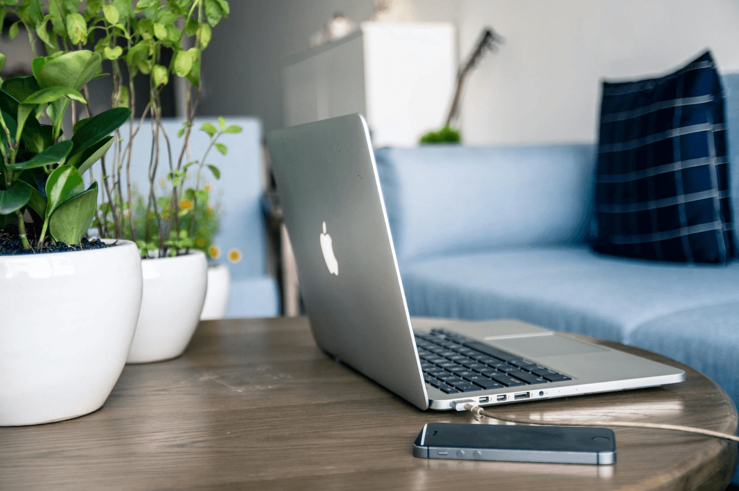 alt="Ordinateur portable ouvert et smartphone posés sur une table basse en bois à côté de plantes d'intérieur, avec un canapé bleu en arrière-plan"