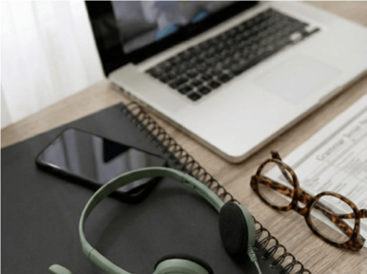alt="Ordinateur portable ouvert sur un bureau affichant une visioconférence, avec un casque audio vert, des lunettes et un carnet au premier plan"