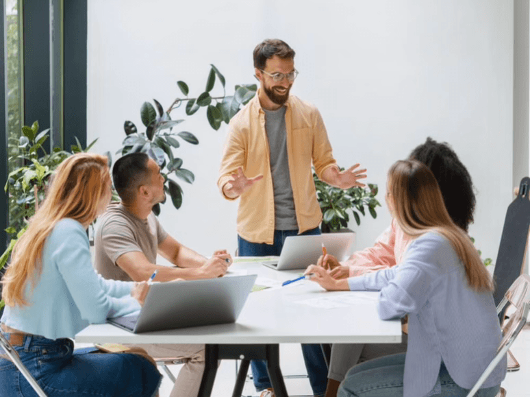 alt="Un homme debout prend la parole devant quatre participants à atelier assis autour d'une table blanche dans un bureau moderne et lumineux décoré de plantes"