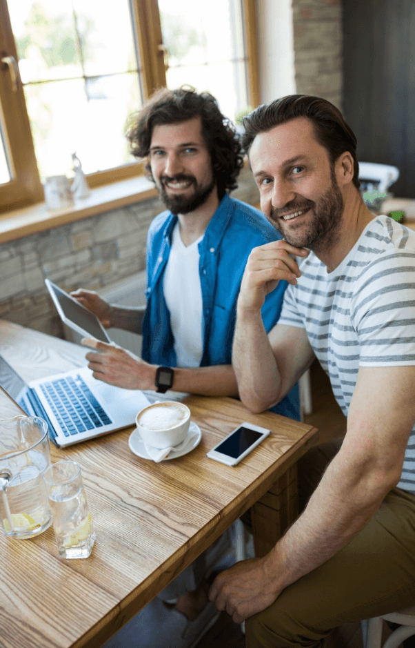 alt="Deux jeunes entrepreneurs sourient à la caméra dans un café. Sur la table, un ordinateur portable, une tablette et un smartphone pour travailler sur leur projet"