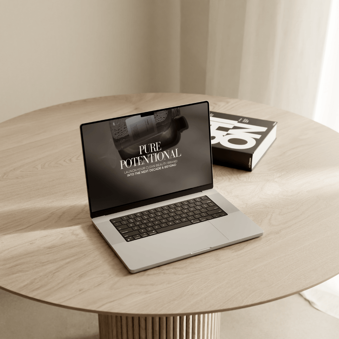 A laptop displaying the text "PURE POTENTIAL" sits on a round wooden table next to a book. The laptop screen also shows "LAUNCH YOUR DREAM BEAUTY BRAND INTO THE NEW DECADE & BEYOND".