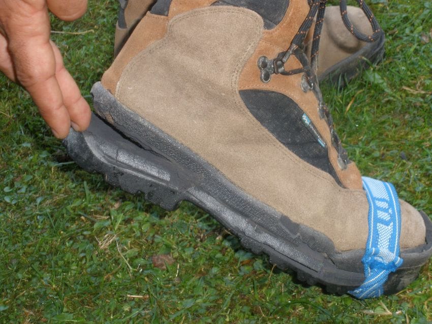 Primer plano de una bota de montaña con la suela despegada, sujeta de forma provisional con una cinta. La bota está sobre un terreno de hierba en la montaña.