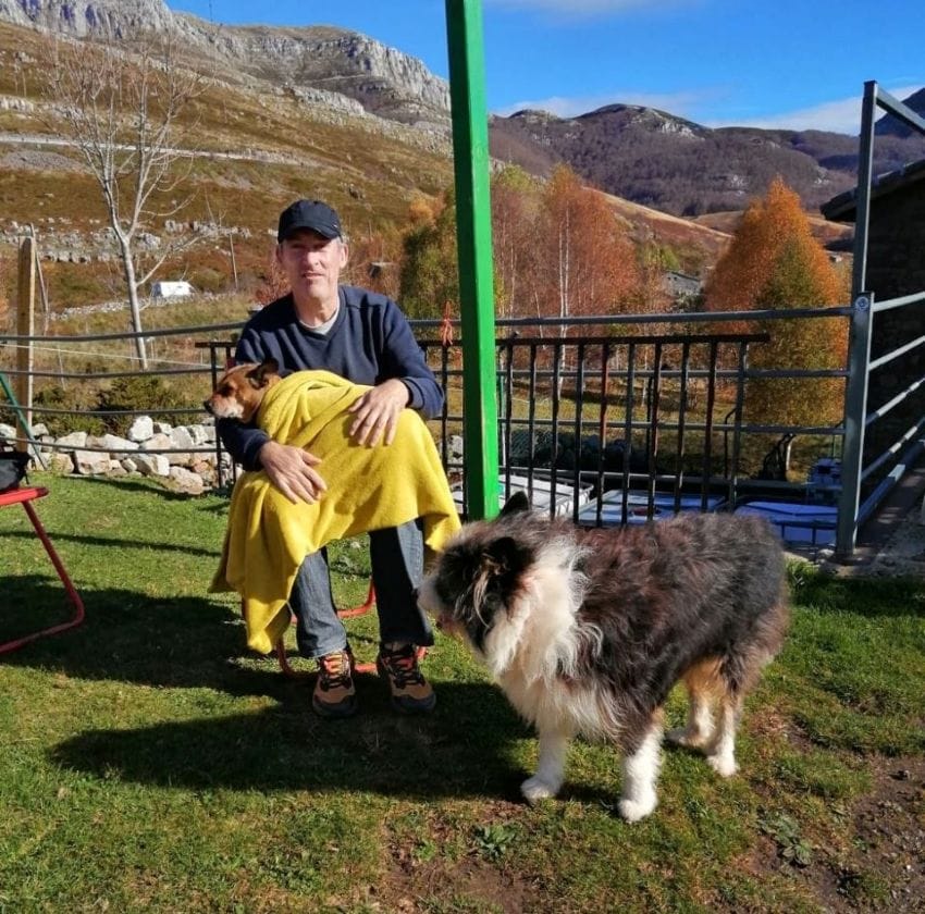 Montañero sentado en un refugio de montaña con su perra descansando sobre sus rodillas bajo una manta amarilla, con picos al fondo.