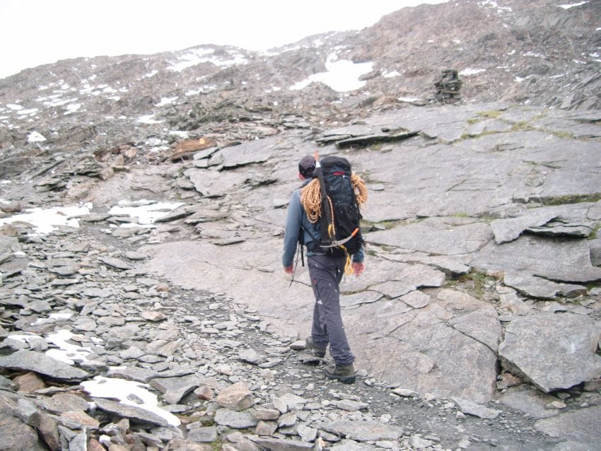 Jesús E. Mamés en un sendero rocoso de alta montaña en los Alpes, cargado con una mochila pesada, piolet y cuerda visible, bajo un cielo blanquecino, ilustrando el esfuerzo sostenido y las señales sutiles de cansancio que el cuerpo empieza a enviar antes de saturarse
