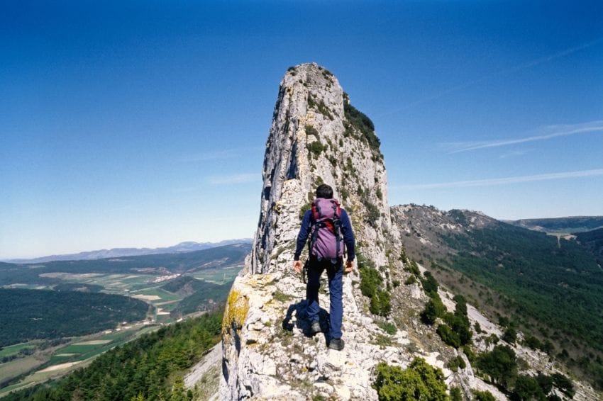 Montañero con una mochila pesada subiendo una cresta rocosa hacia una gran torre, ilustrando el peso acumulado del estrés mental.