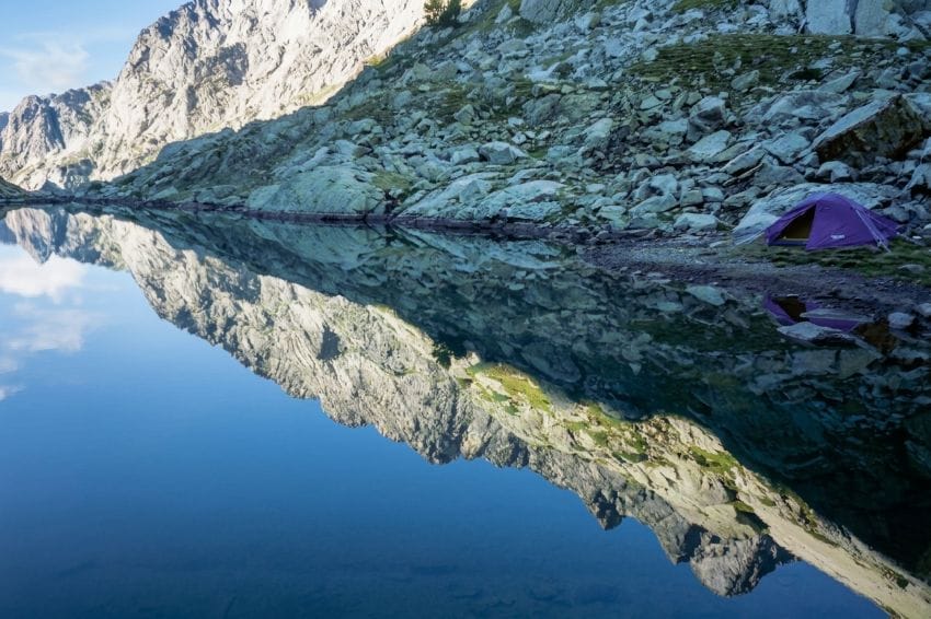 Tienda de campaña junto a un lago de montaña reflejando el paisaje en calma para ilustrar la necesidad de liberar carga mental