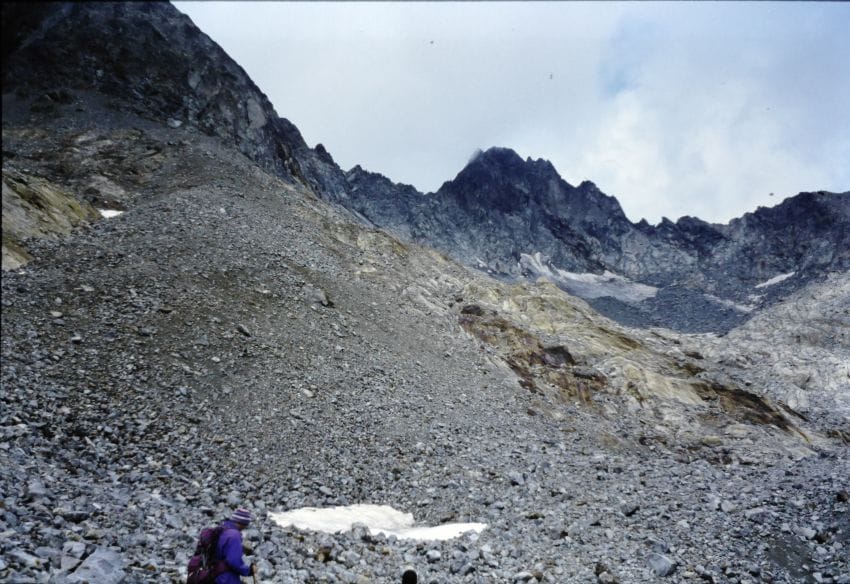 Montañero caminando por un terreno de rocas y pedregal empinado hacia la cumbre sufriendo desgaste físico.