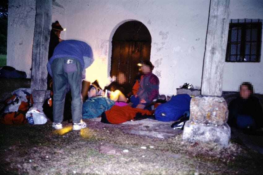Grupo de montañeros descansando en el suelo de un pórtico de ermita tras una larga jornada de actividad, mostrando el cansancio y la postura de descarga.