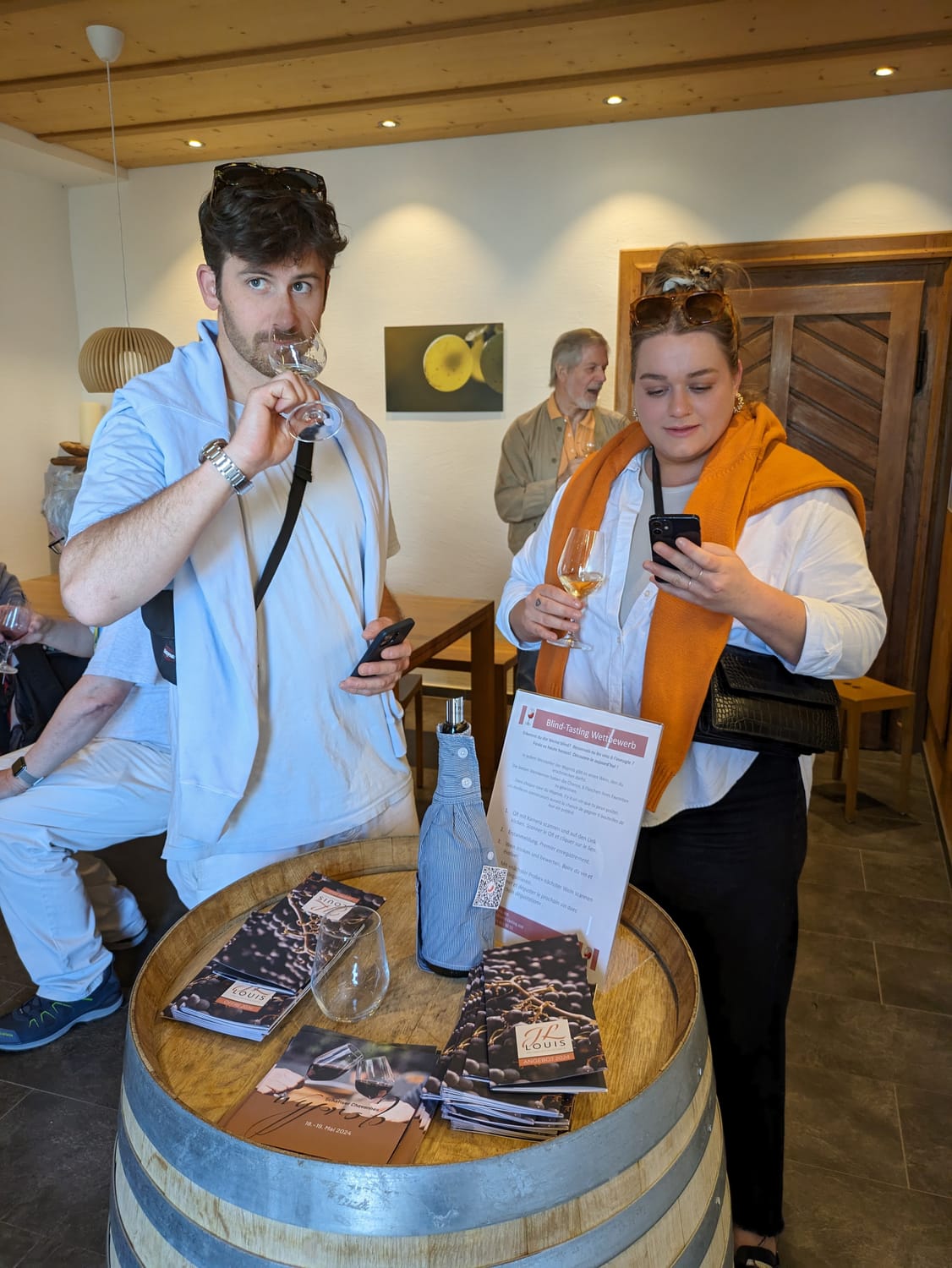 In einem kellerartigen Raum stehen zwei Personen die an der Blindverkostung mitmachen. Sie verwendet ein Smartphone um den auf der blinden Weinfalsche angebrachten QR-Code zu scannen. Im Hintergrund sind Holzfässer zur sehen.