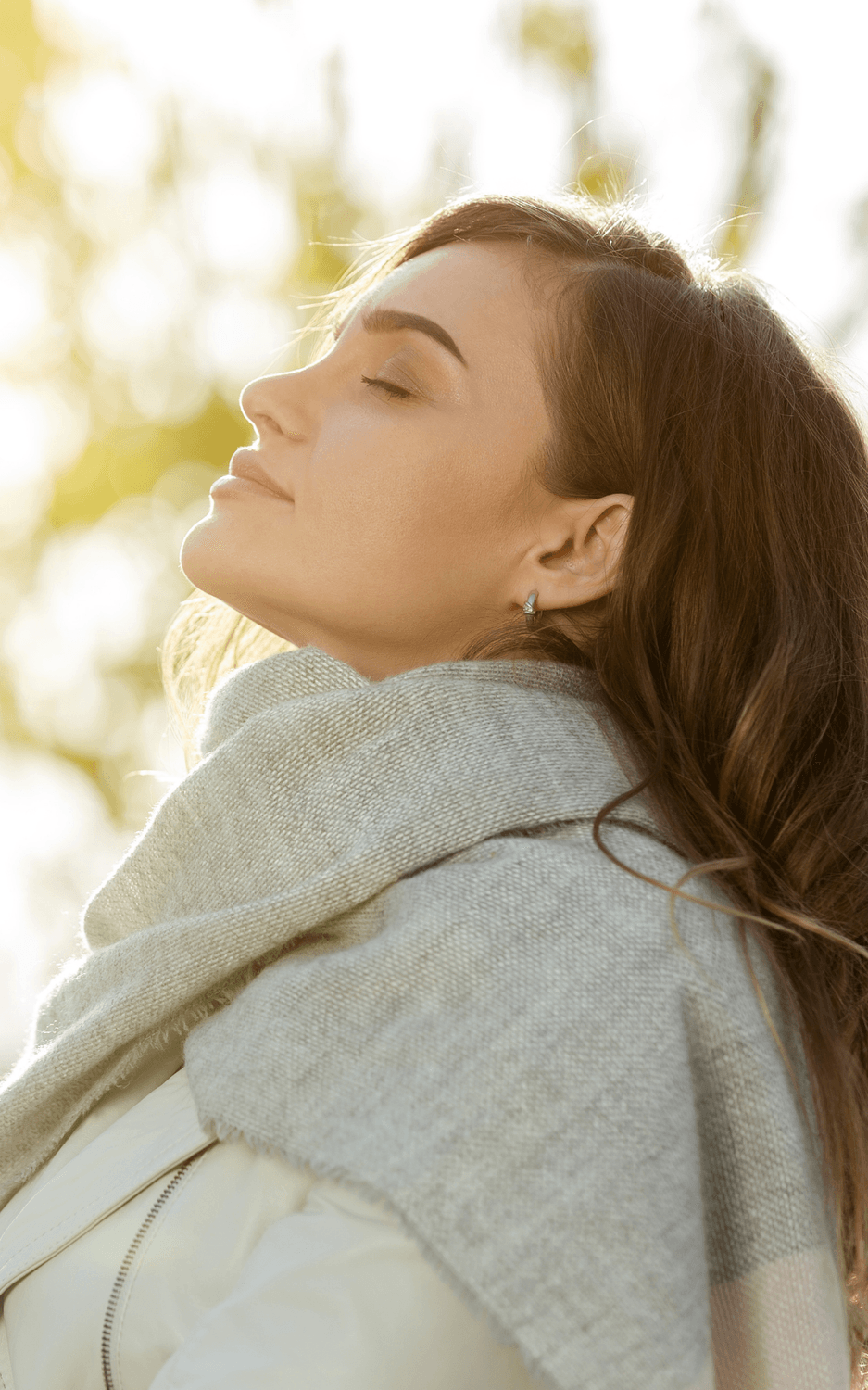 Nainen seisoo ulkona silmät suljettuina ja hengittää rauhallisesti syvään, kasvoillaan tyytyväinen ilme. Kuva välittää rauhan, palautumisen ja hyvinvoinnin tunnetta.