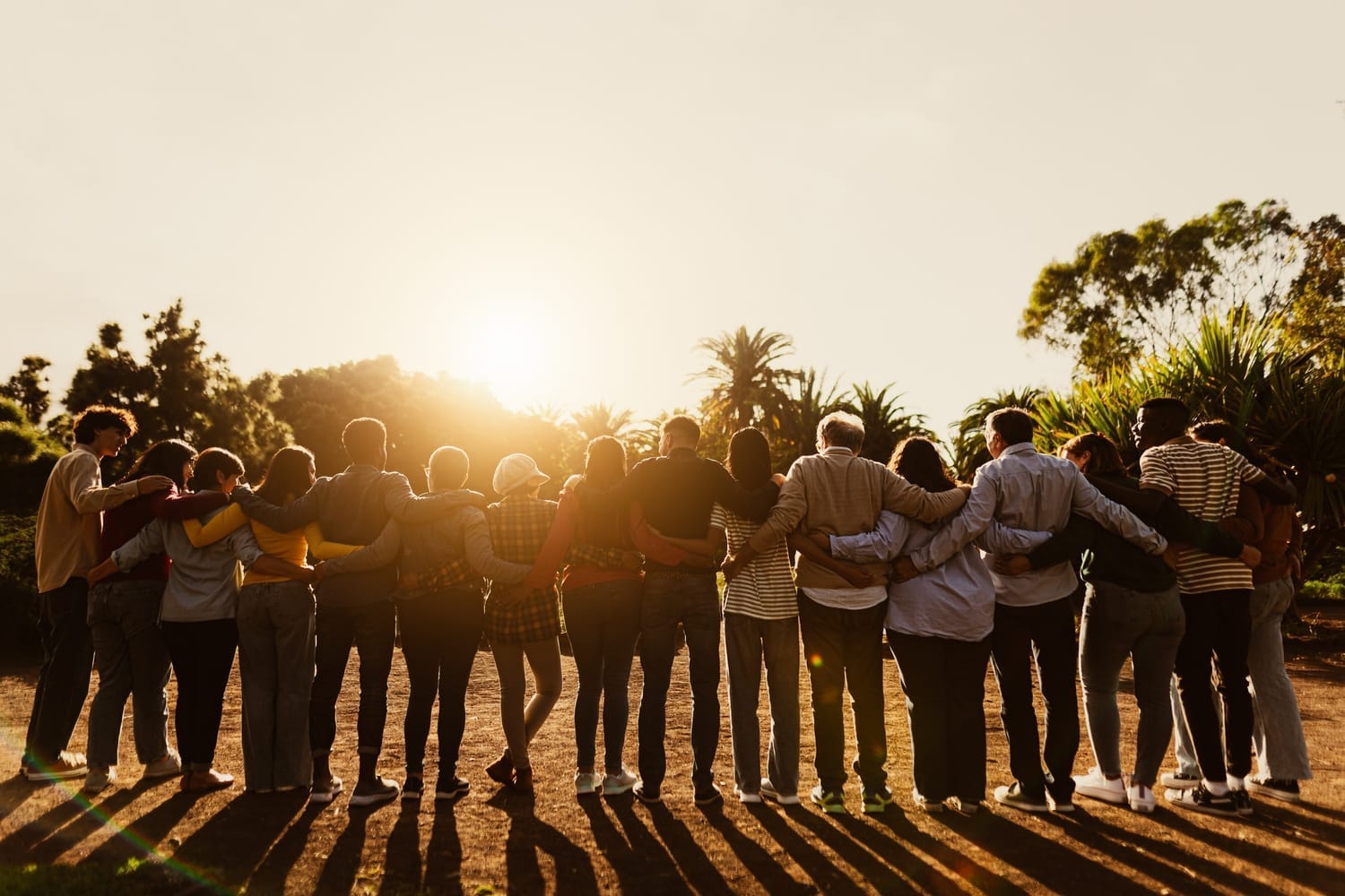 Group of 16 people standing side by side with their backs to the camera, watching the sunset, each person holding the person next to them around the back, symbolizing unity and empathy for others