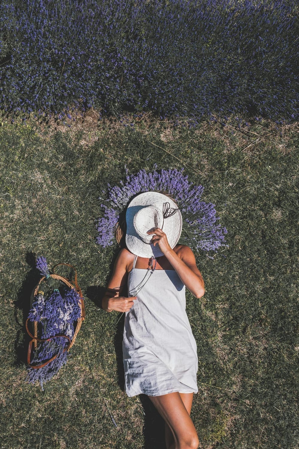 Woman lying on grass in a white summer dress, covering her face with a white hat, with a basket of lavender beside her and lavender arranged like sun rays around her head, symbolizing self-empathy and peaceful self-connection