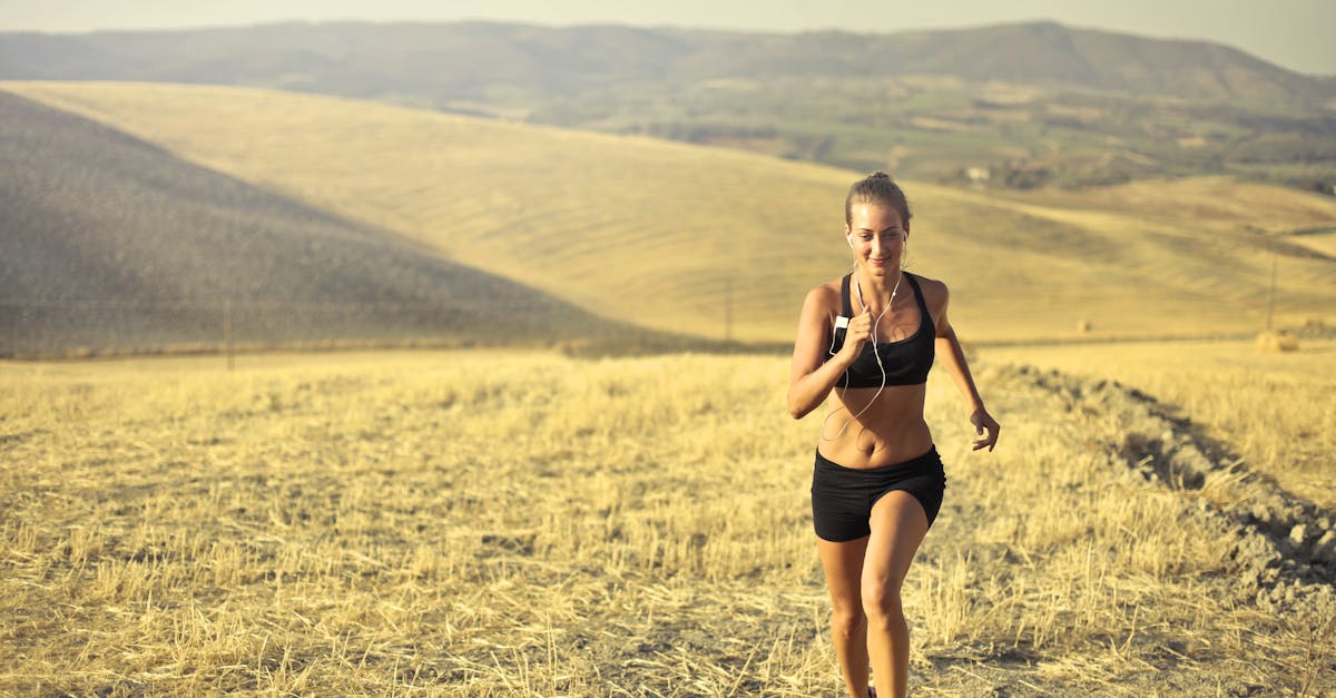 Powerful young female athlete in activewear running along hill on background of mountainous landscape and listening to music in earphones during cardio training