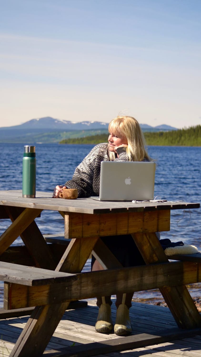 Pernilla Öberg sitting with her MacBook by a calm lake in the Swedish arctic mountains of Vallrun