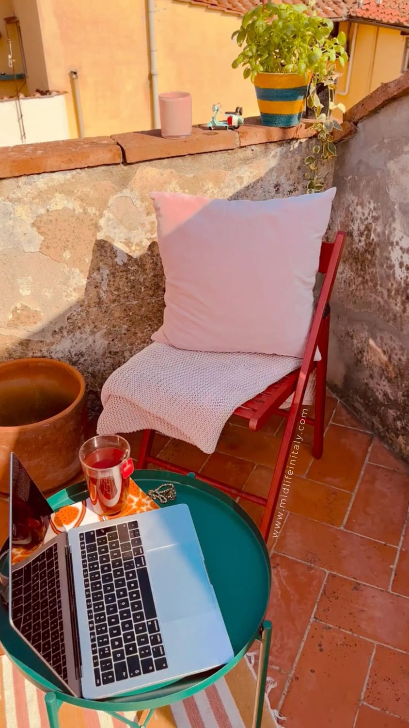 A midlife woman working on a laptop on a sunny rooftop in Florence, Italy, building an automated income system from her Handbag Office.