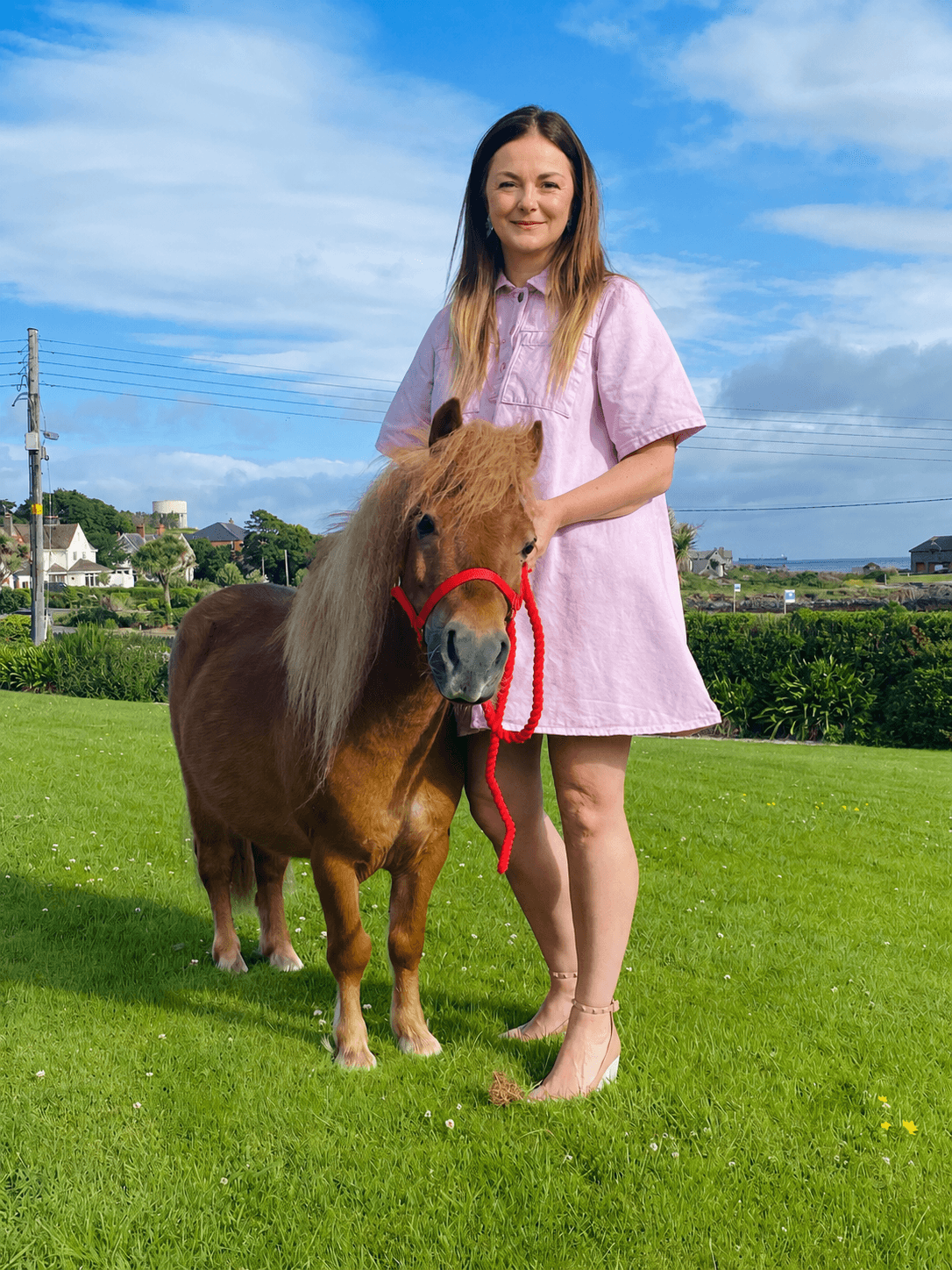 A young woman standing in her garden with a lilac denim dress on. She stands beside a chestnut miniature Shetland pony with a red harness on