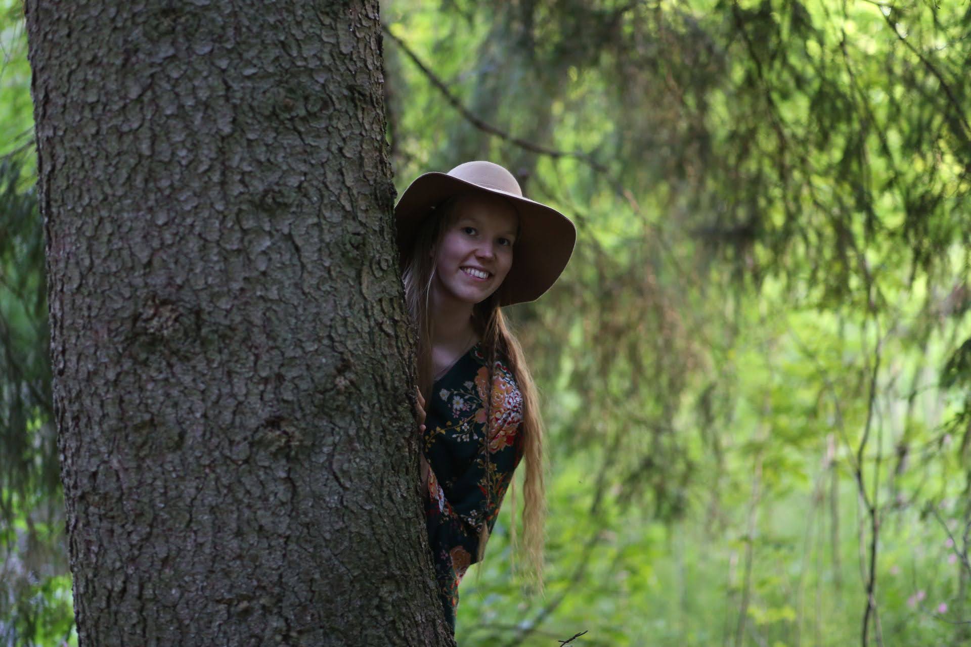 Nainen jolla hattu päässä kurkistaa puun takaa