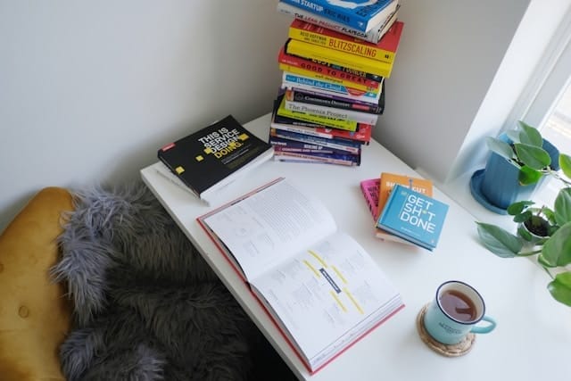 Stack of books on a variety of topics on desk beside a cup of coffee.