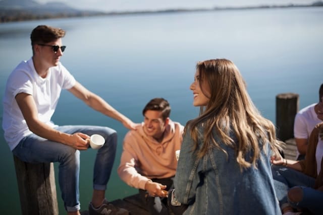 Friends seated on a dock on a lake enjoying cups of coffee.