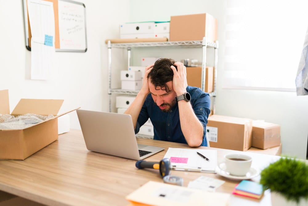 Stressed online store owner holding his head at a laptop surrounded by shipping boxes, illustrating the cost of unreliable ecommerce website hosting