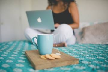 Una taza celeste con galletas sobre una tabla de madera, y Marianella Ruiz sentada en la cama con su computador, al fondo