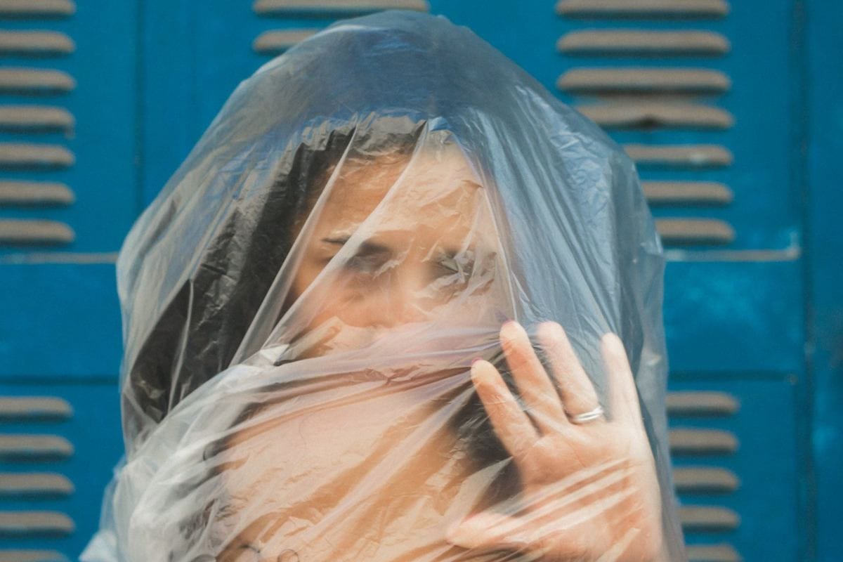 Marianella Ruiz con el rostro cubierto por un plástico transparente y una mano al frente, frente a un portón azul