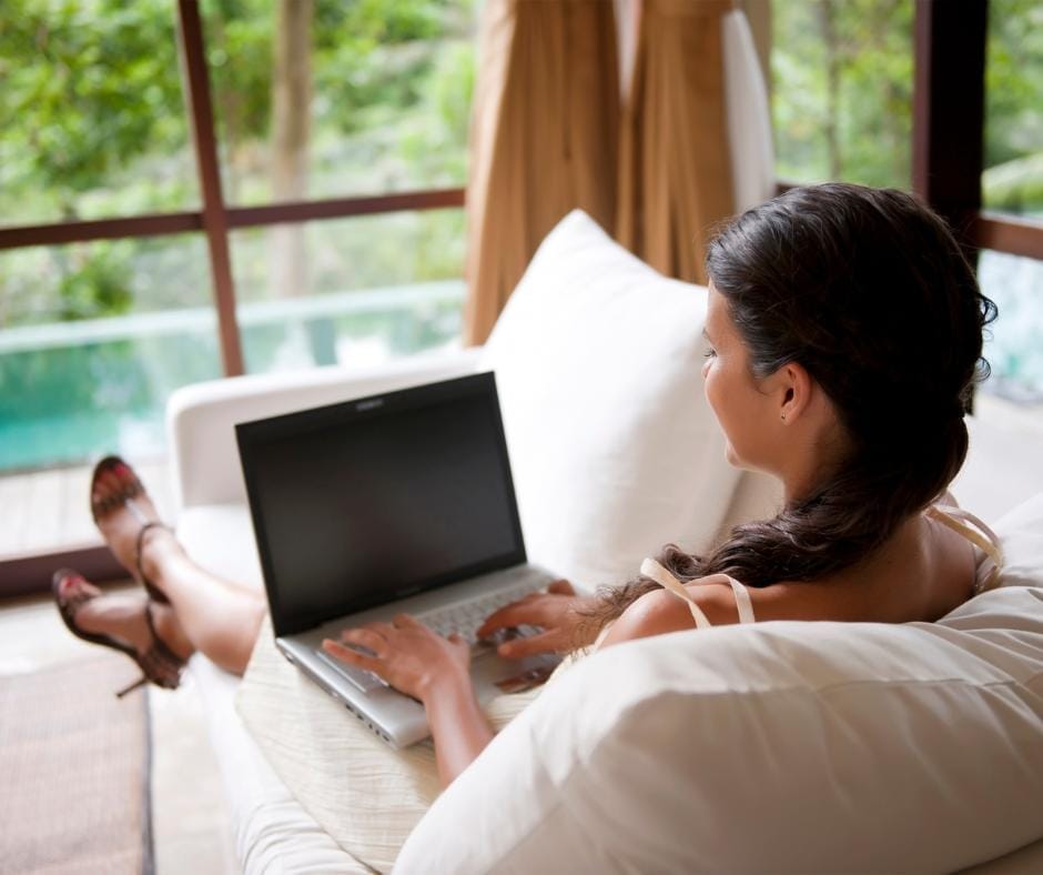 Woman working from home sitting on her couch using her laptop to work relaxing infront of a nice outside view