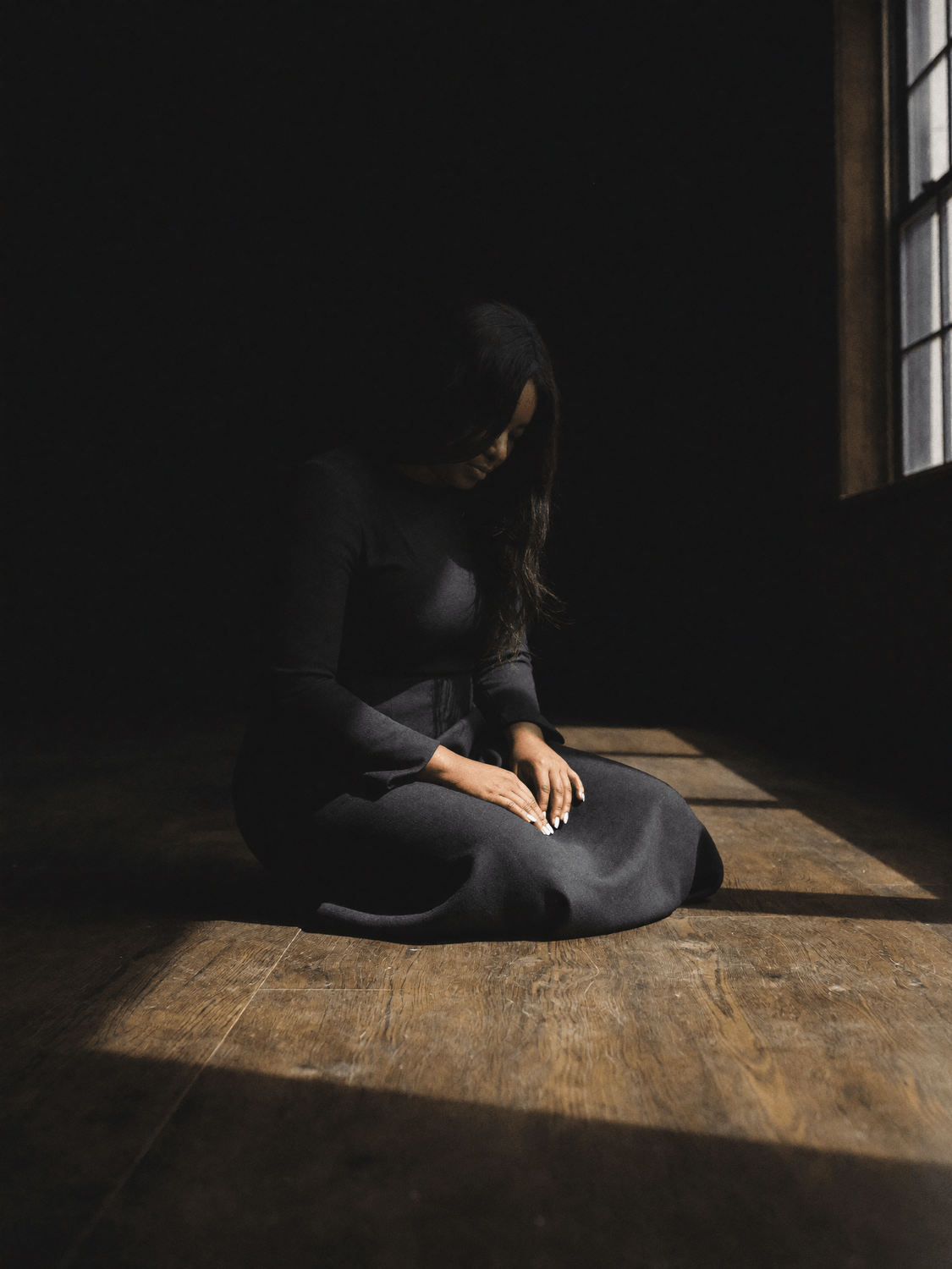 Woman sitting alone in dark room with single shaft of golden light, depicting spiritual emptiness and feeling distant from God