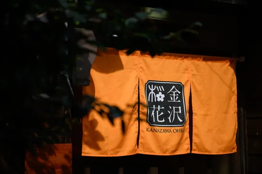 an orange noren (a traditional Japanese fabric divider) hanging in front of a dark entrance. The noren features Japanese characters and the text "KANAZAWA OH," with a stylized design above the text. The area surrounding the noren appears shaded, with some greenery partially visible in the foreground.