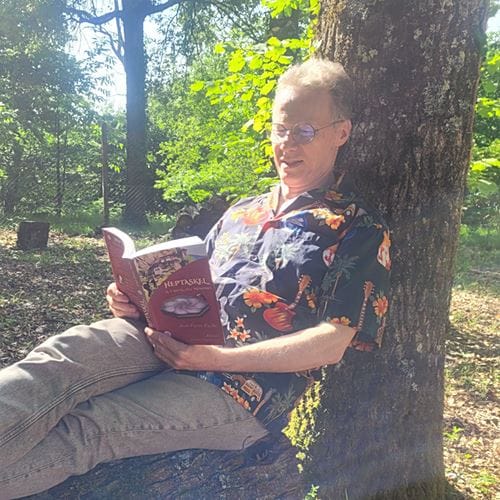 Photo de l'auteur (Jean-Pierre Fuchs) adossé à un arbre et lisant son premier roman "Heptaskel - Le Cristal des Mémoires"